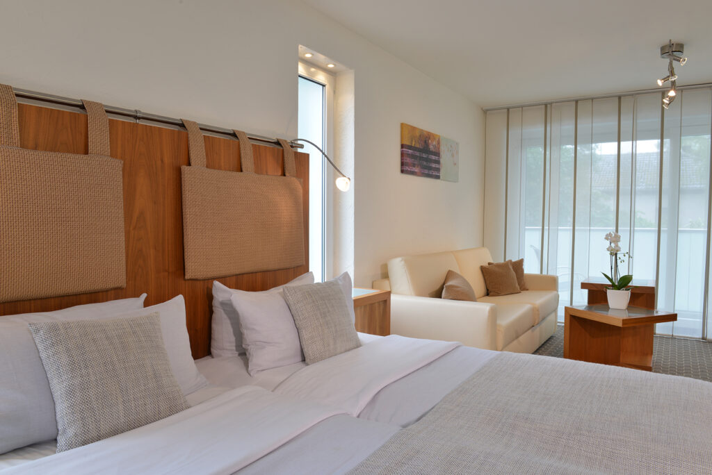 Deluxezimmer Parkhotel Oberhausen Blick seitlich auf einen Teil des Bettes. Im Hintergrund eine Sitzecke. beige und hellgrau