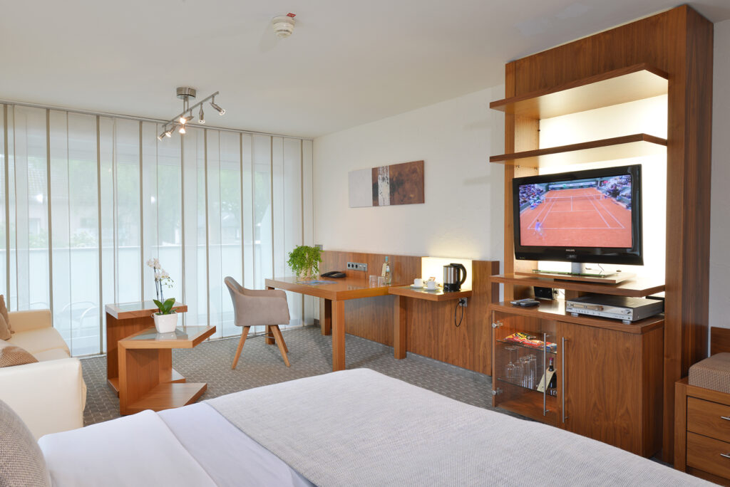 Deluxezimmer Parkhotel Oberhausen Blick seitlich auf einen Teil des Bettes. Im Hintergrund TV Wand und Schreibtisch. beige und hellgrau