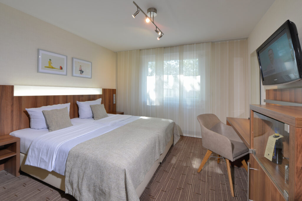 Komfortzimmer Parkhotel Oberhausen Blick in das Zimmer auf das Doppelbett und rechts mit Schreibtisch und TV an der Wand. in braun und grau T&ouml;nen gehalten.