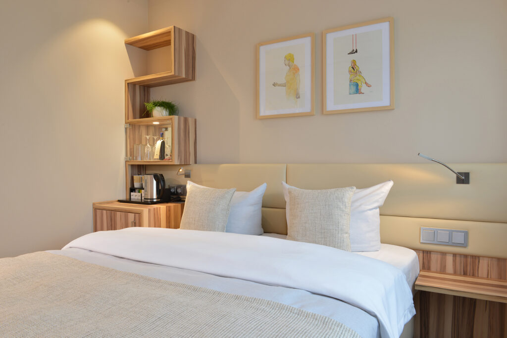 Standardzimmer im Parkhotel Oberhausen mit Blick auf das Bett an der Wand mit 2 Bildern und einem Regal. in beige gehalten