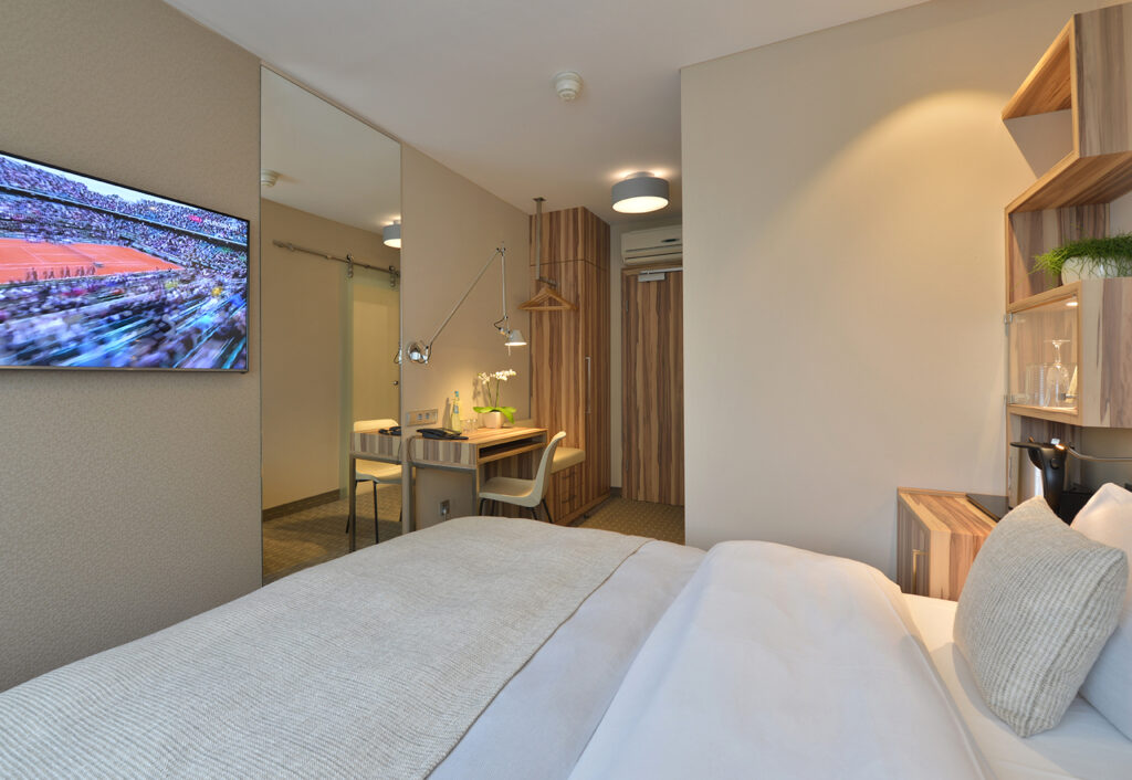 Standardzimmer im Parkhotel Oberhausen mit Blick zur Eingangst&uuml;r. Im Vordergrund ein Teil vom Bett, TV an der Wand gegen&uuml;ber. In beige gehalten.