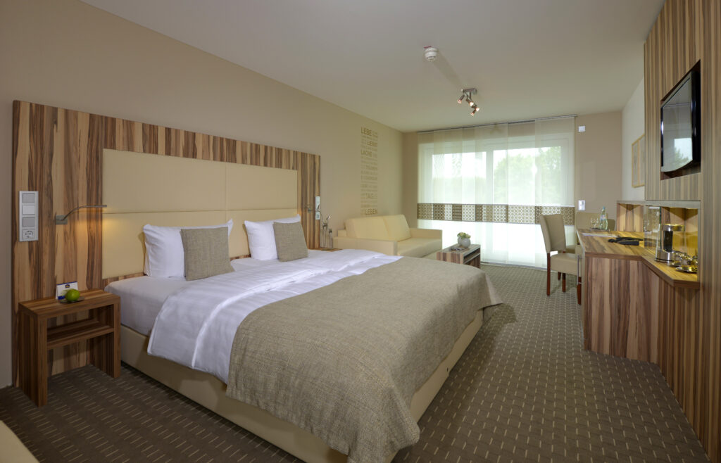 Juniorsuite Zimmeransicht mit Doppelbett, Schreibtisch und Fensterfront. Warme beige und braunt&ouml;ne