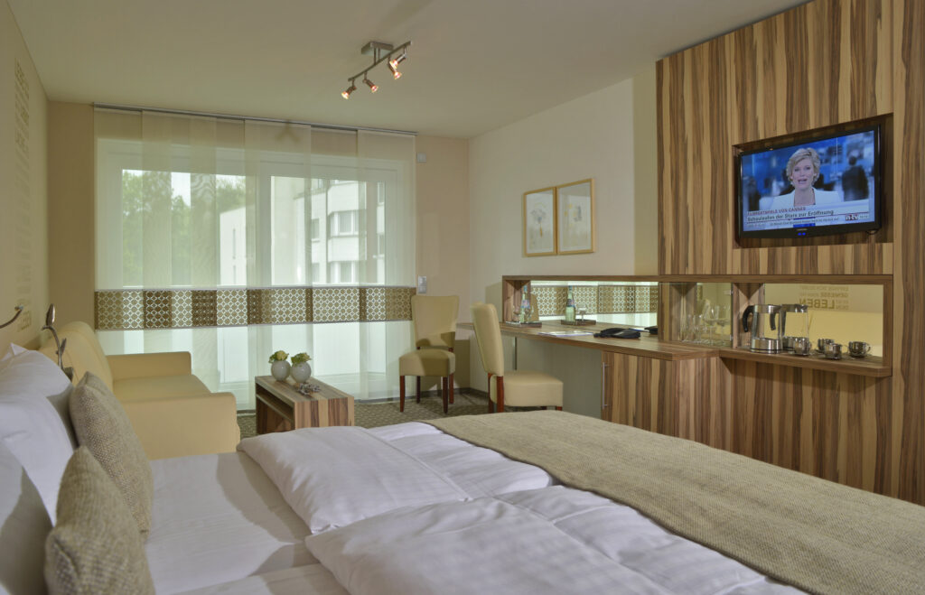 Juniorsuite Ansicht Bett auf Schreibtischwand mit Spiegel und Wasserkocher. Links ist ein beiges Sofa hinter dem Bett zu sehen.