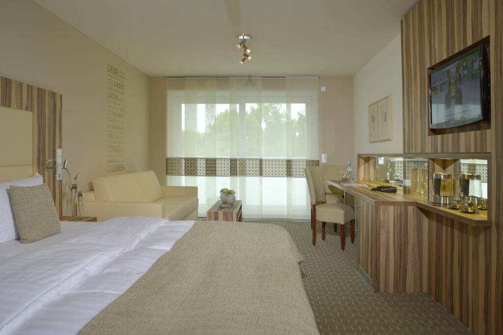 Juniorsuite. Gerader Blick ins Zimmer mit Sicht auf Bett, Schreibtisch, Sitzecke und Fensterfront. warme beige und braun T&ouml;ne