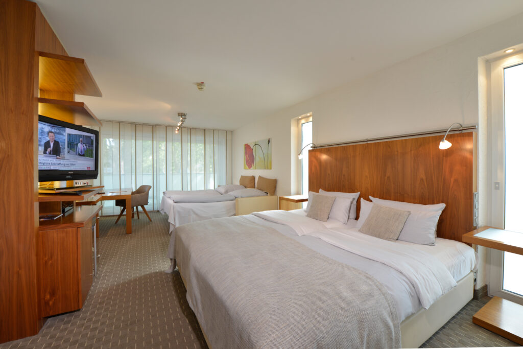 Deluxezimmer Parkhotel Oberhausen als Familienzimmer. Blick auf das Doppelbett, im Hintergrund die aufgebette Schlafcouch. beige und hellgrau gestaltet