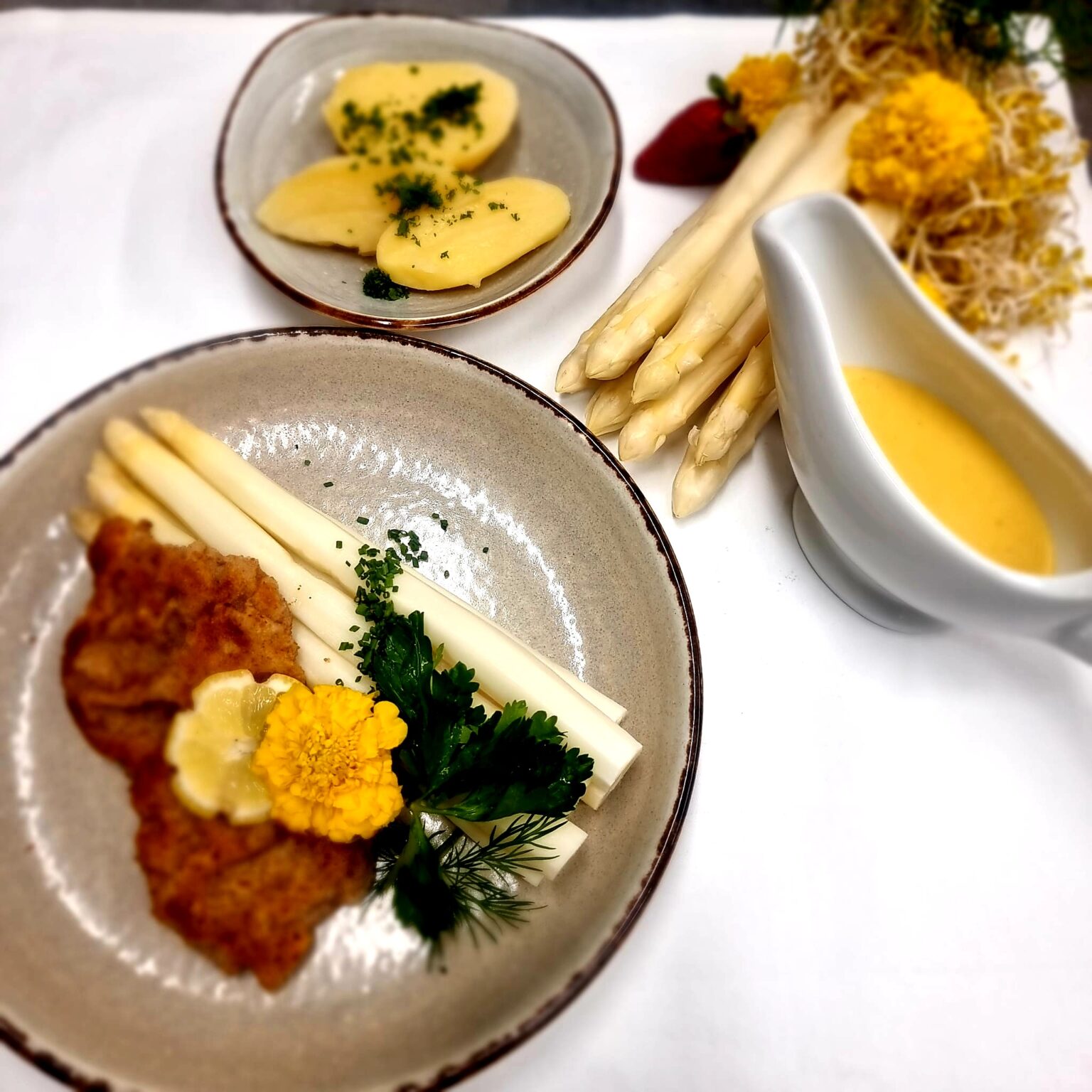 Teller mit Spargel, Schnitzel. Daneben ein Teller mit Kartoffeln eine weiße Sauciere mit Sauce Hollandaise und noch Stangenspargel als Deko